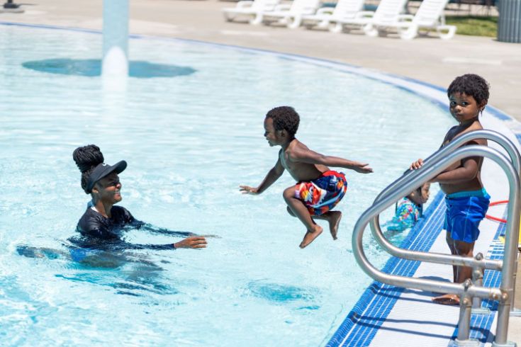 Mother and two kids at public pool. One child jumping into the water and smiling.