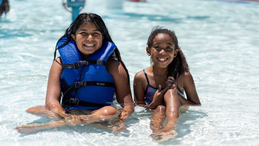 Two kids in a pool smiling for the camera, one wearing a life vest