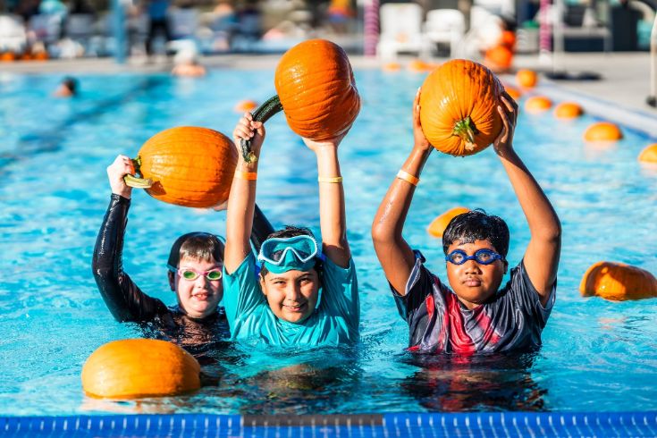 Three kids in a pool holding pumpkins over their heads