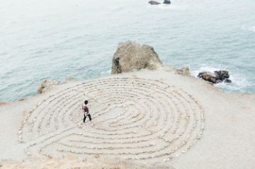 Person walking through sand maze with ocean and large rocks in background.