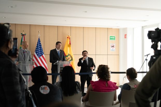 Three men stand at a clear podium during a press conference, flanked by the American flag and what appears to be the flag of Morelia. Media cameras are visible in the foreground, and an audience is seated facing the speakers.