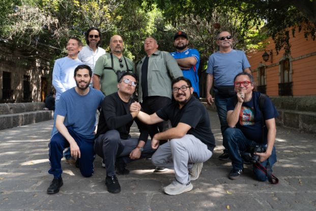 A group of approximately ten men pose for a casual photo outdoors in a tree-shaded plaza next to a pink stone colonial building. Some are crouching in front while others stand behind.