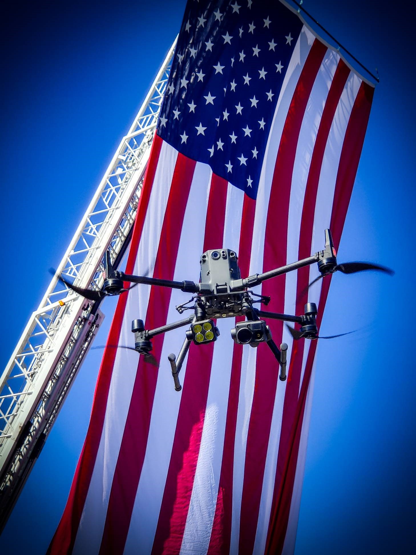 A photo of an uncrewed aircraft system hovering in front of an American flag mounted on the top of a fire truck's extended ladder.