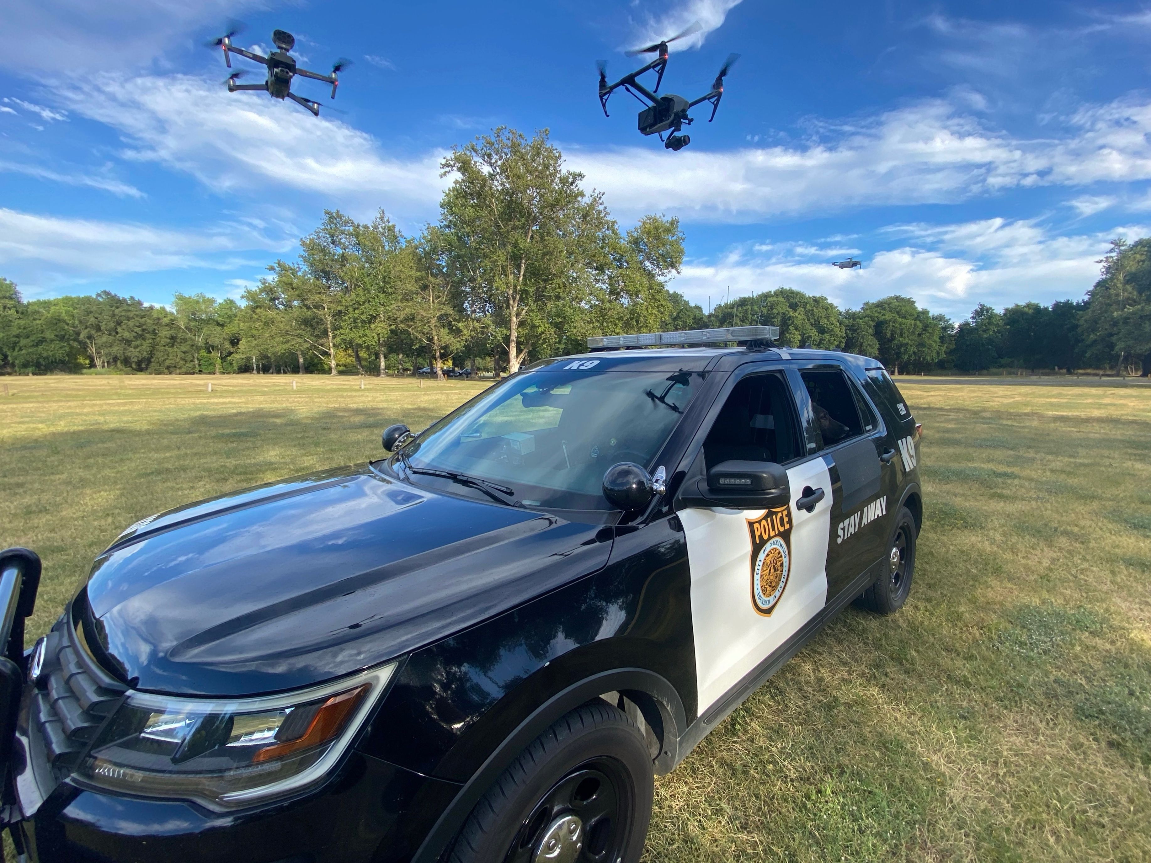 A photo of a fully-marked Sacramento Police Department K9 patrol vehicle parked in a field with two police operated unmanned aircraft systems flying overhead.