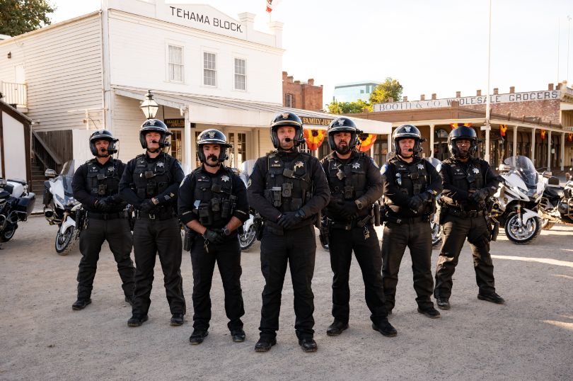 Seven motor officers with their motorcycles behind them in front of a building in old sac
