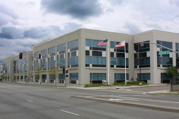A photo of the Sacramento Police Department's Richards Police Facility (Central and East Commands) . It is a grey building with blue tinted windows facing Richards Boulevard. The National and State flags can be seen on display out front.