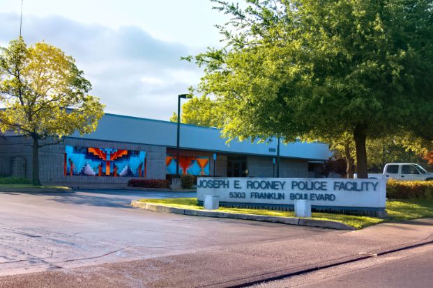 A photo of the Sacramento Police Department's south area Joseph E. Rooney Police Facility (Central and East Commands) . It is a grey building with white trim and a colorful mural on the front wall. There are also trees, grass and a sign with the building's name out front.
