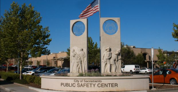 A photo of the Public Safety Admin Building, which houses the headquarters for the Police and Fire Departments. It is a beige building with a parking lot and statue in the foreground. Cars and trees are visible in the parking lot, as is a flag pole with the US flag