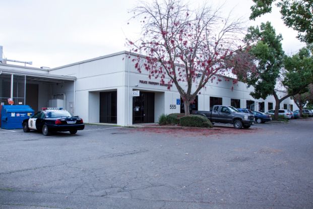 a photo of the Sacramento Police Department Evidence and Property building. It is a white building, and there is a police vehcile parked out in front. The parking lot also has several trees and bushes.