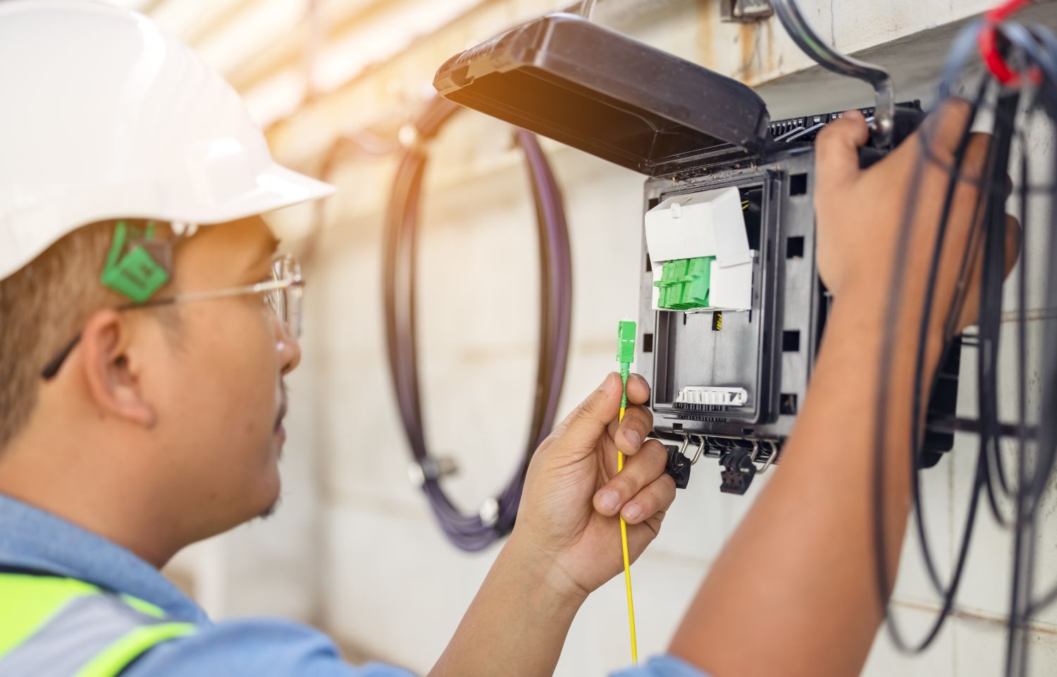 A utility worker in a hardhat, glasses, and high-visibility vest holds up a fiber optic internet cable near an open internet connector box.  