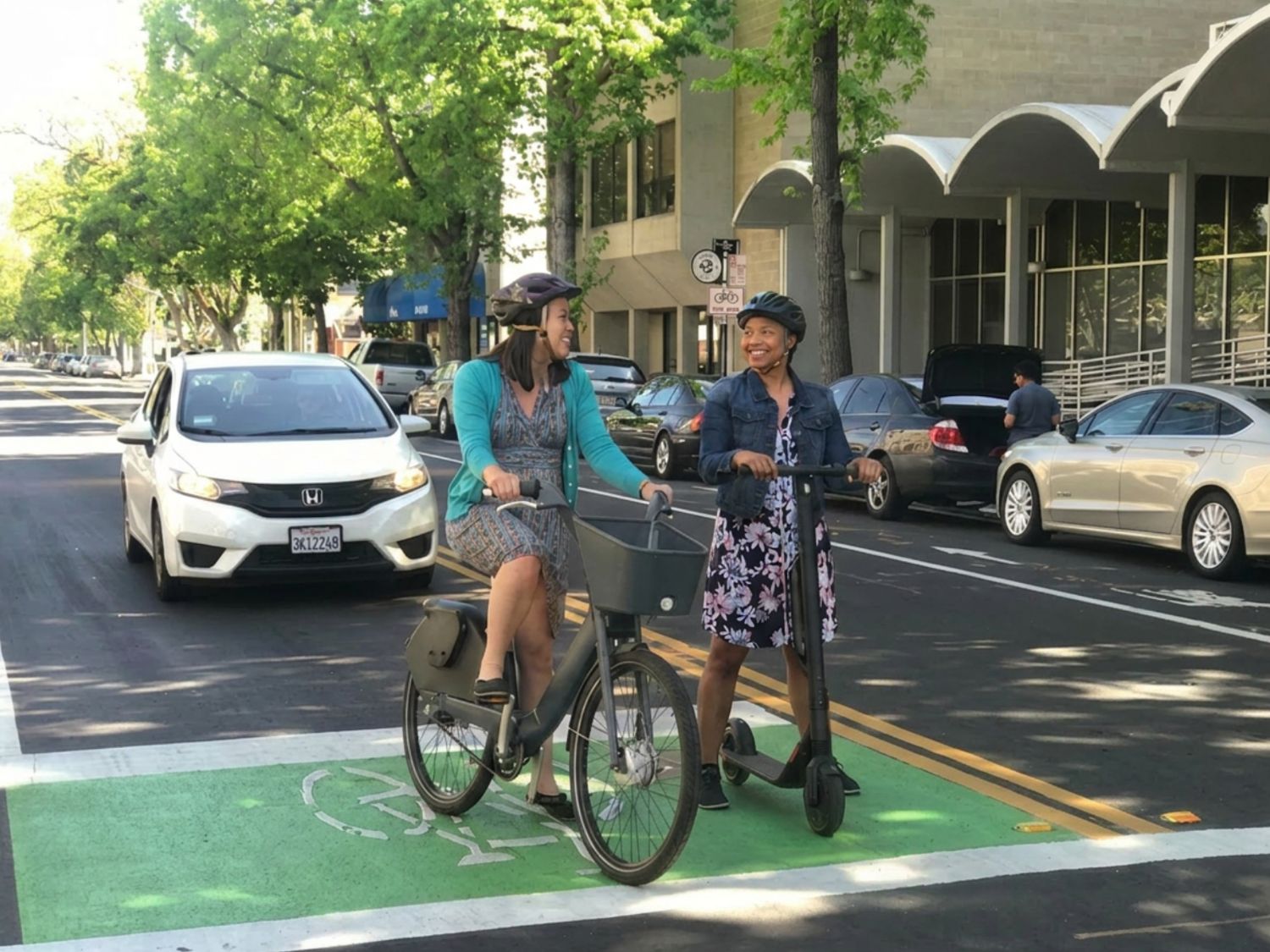 A person on a bike and a person on a scooter in a bike box on a tree-lined street. 