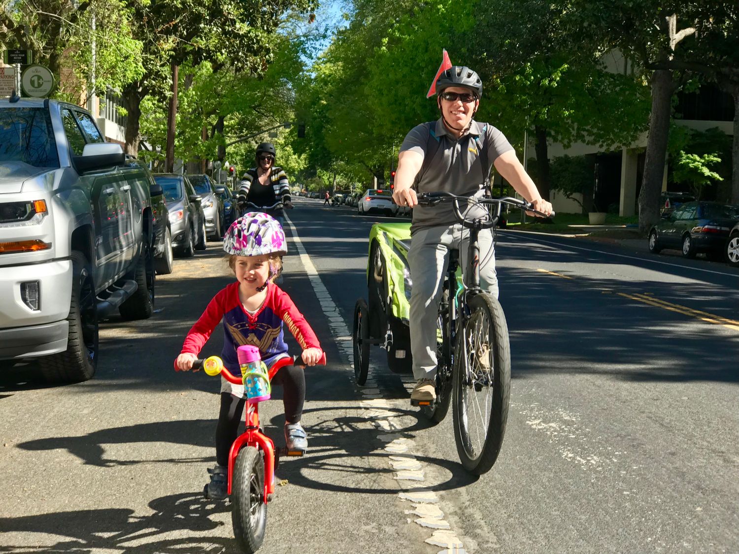 Image of a child on a red bike accompanied by a parent on an e-bike towing a trailer and a parent riding behind on a bicycle.